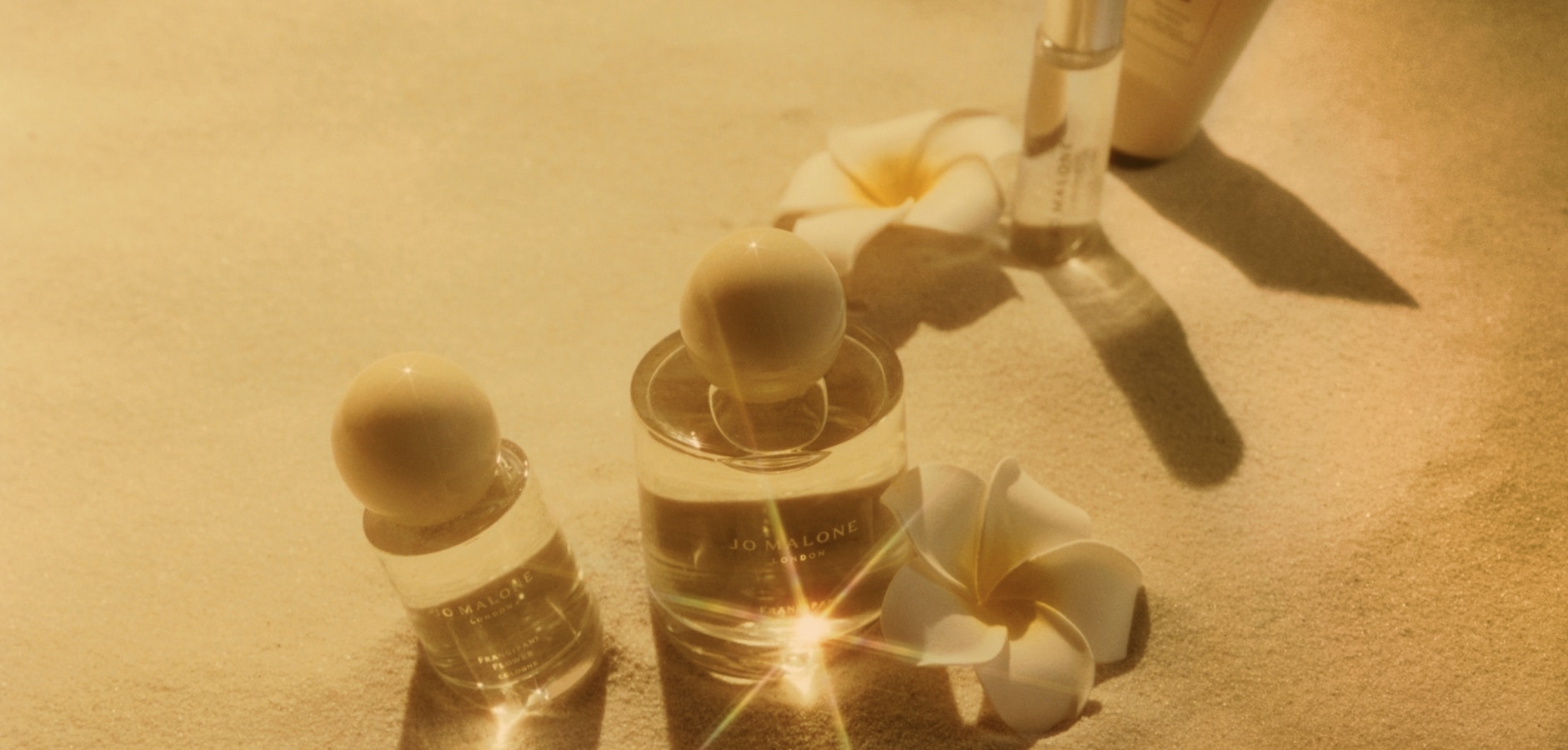 A Jo Malone London cologne rests atop a sandy beach then pans to a person holding it in white swimwear. 