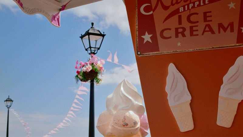 A person in pink takes an icecream then walks along a pier containing vibrant beach props before dropping the icecream.