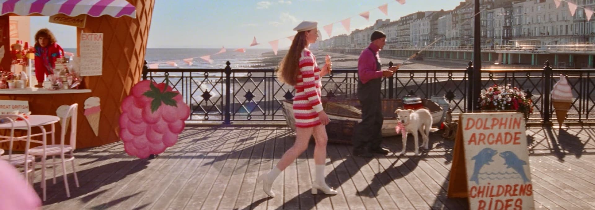 A person holds an icecream whilst walking along a pier full of people and vibrant beach props before dropping the icecream, 
