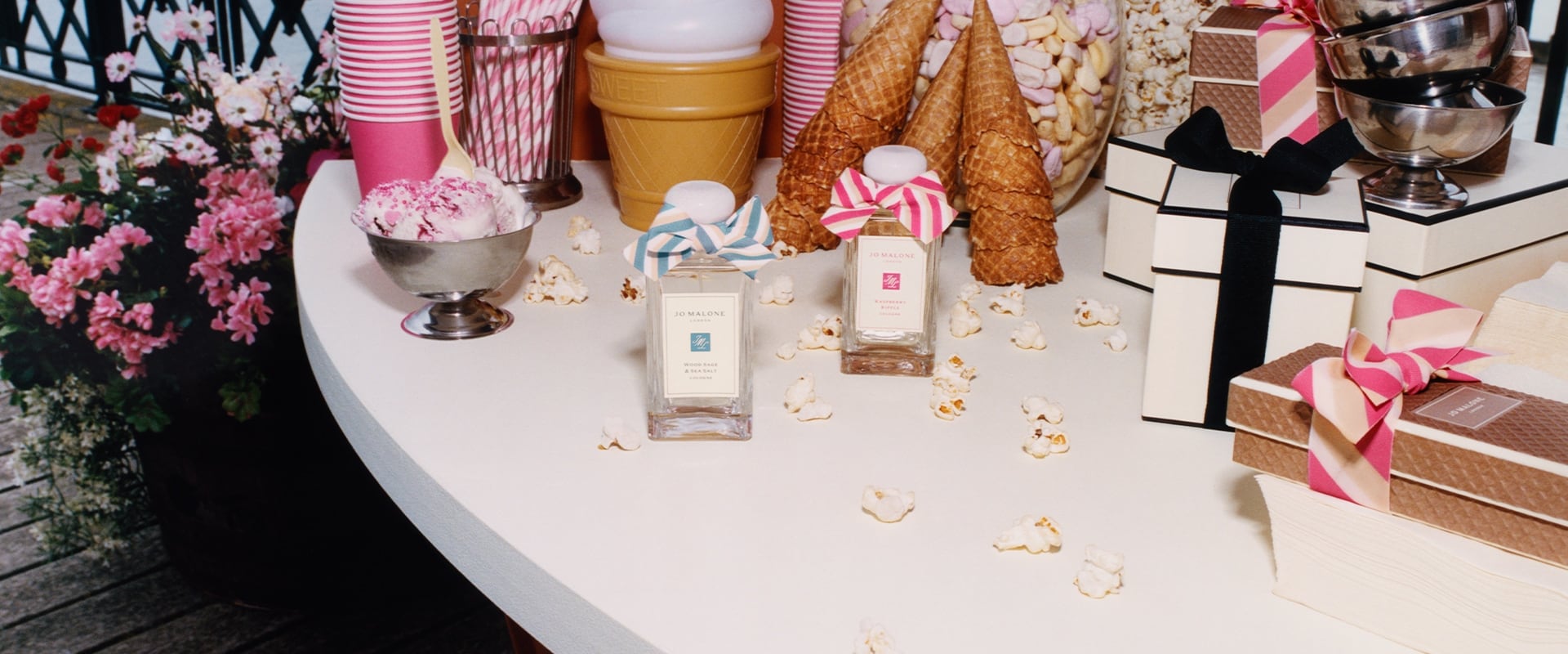 Two Jo Malone London colognes, one with a blue bow, one with a pink bow surrounded by pink cups and straws, popcorn and sweets.