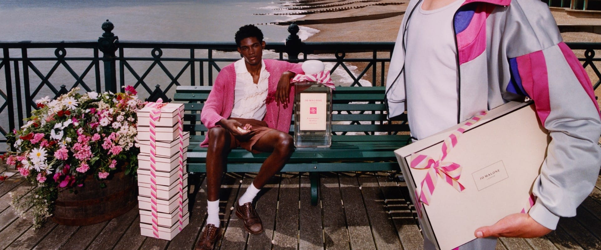A person holding an oversized icecream walks past a person on a bench next to an oversized cologne beside a stack of gift boxes.